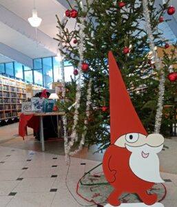 Kuvassa punaiseen pukeutunut puinen tonttu valkoisella parralla on joulukuusen edessä, takana kirjahyllyjä ja pöytä täynnä kirjoja.